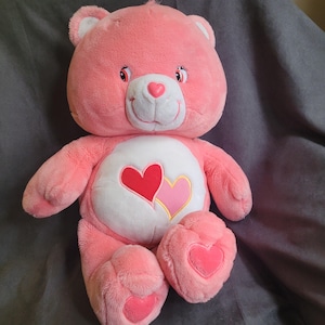 Puede incluir: Un oso de peluche rosa con una barriga blanca con dos corazones. El oso tiene una cara sonriente con una nariz rosa en forma de corazón y acentos en forma de corazón en sus patas. Un juguete suave y tierno.