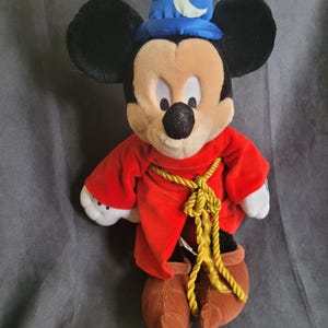 May include: A plush Mickey Mouse toy dressed as a sorcerer. The toy has black ears, a tan face, and is wearing a red robe with a gold rope belt and a blue hat with a crescent moon. The toy's shoes are brown.