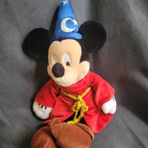 May include: A plush Mickey Mouse dressed as a sorcerer, wearing a blue hat with a crescent moon and star, a red robe, and brown boots. The toy has black ears, a black nose, and white gloves. A yellow rope is tied around the robe.