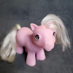 Puede incluir: Un juguete My Little Pony rosa con una melena y cola blancas. El pony tiene sombra de ojos morada y ojos grandes y oscuros.