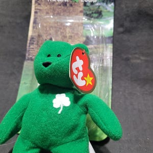 May include: A green plush bear named Erin, with a white shamrock on its chest. The bear has a red tag with a star and the Ty logo. The packaging behind the bear has the name "Erin" and the words "The Bear Yours".