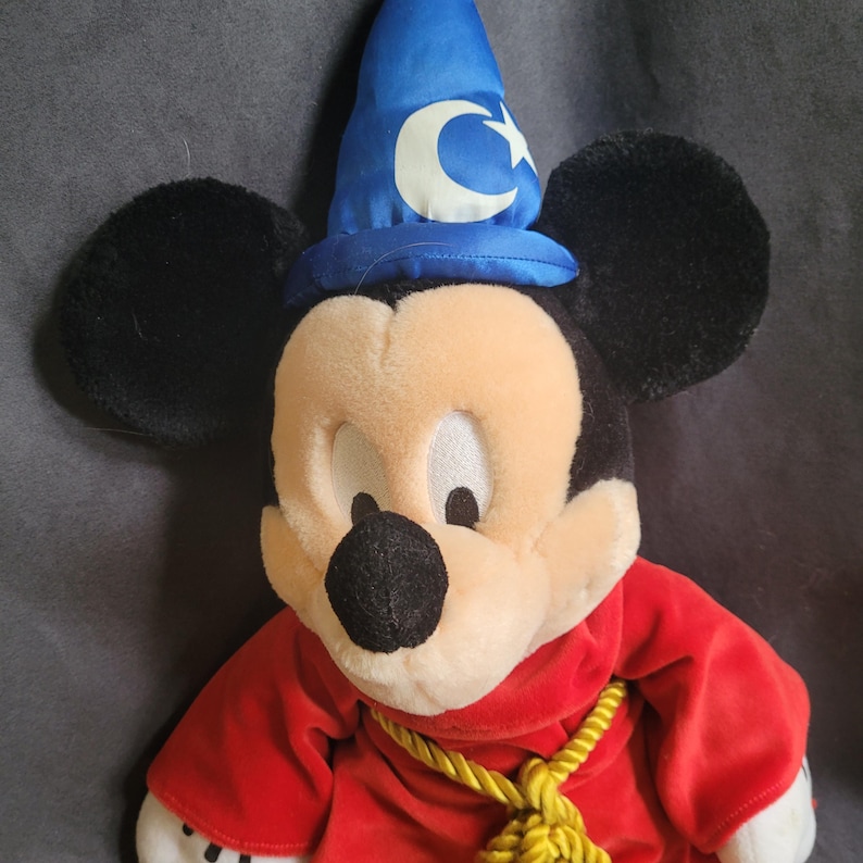 May include: A plush Mickey Mouse dressed as a sorcerer, wearing a blue hat with a white crescent moon and star. He has a red robe and a gold rope around his neck. The plush toy has black ears and a black nose.