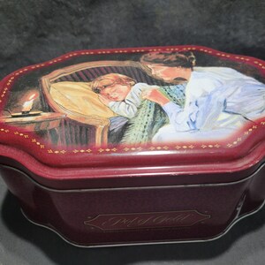 May include: A burgundy-coloured, octagonal tin with a lid featuring a painted scene of a child sleeping in a bed, with a woman watching over them. A lit candle sits on a table. The words "Pot of Gold" are printed on the tin.