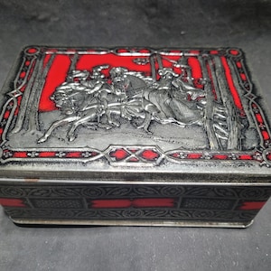 May include: A rectangular metal box with a hinged lid. The lid features a red background with a silver relief scene of figures on horseback. The box has a silver finish with red accents and decorative patterns.