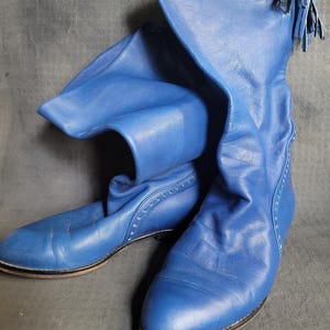 以下が含まれることがあります： 先のとがったつま先と、片方のブーツの上部にタッセルが付いた、青い革のニーハイブーツのペアです。
