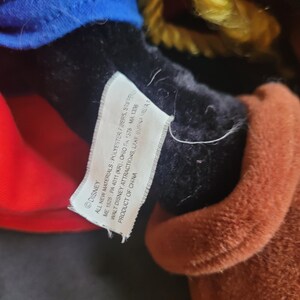 May include: Close-up of a plush toy with a tag. The toy has a blue hat, red shirt, and brown fur. The tag reads "DISNEY" and includes details about materials and manufacturing.