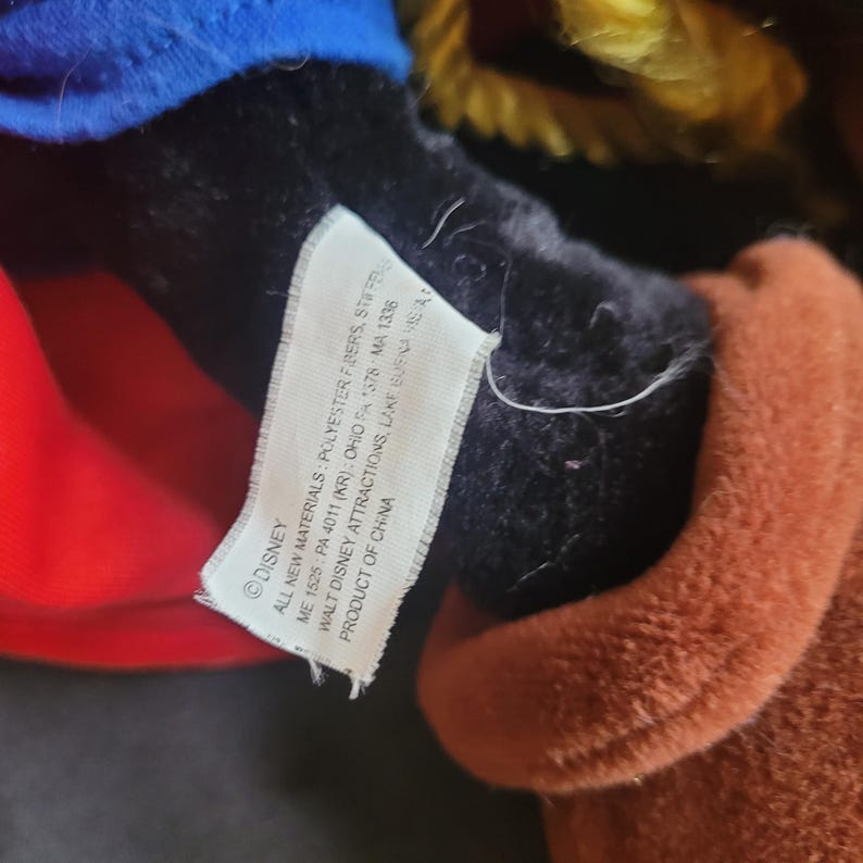 May include: Close-up of a plush toy with a tag. The toy has a blue hat, red shirt, and brown fur. The tag reads "DISNEY" and includes details about materials and manufacturing.
