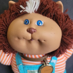 May include: A vintage Cabbage Patch Kids doll with a tan face, blue eyes, and brown hair. The doll is wearing a blue overall with orange and white striped sleeves. A brown collar and a gold-colored tag are around the neck.