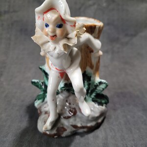 May include: A white porcelain figurine of a whimsical character with a red and white hat, blue eyes, and a red and white outfit. The figurine is holding onto a brown tree trunk and is standing on a base with green leaves.
