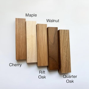 Puede incluir: Una colección de seis muestras de madera rectangulares, cada una mostrando un tipo de madera diferente: Cerezo, Arce, Roble Rift, Nogal y Roble Quarter. Las muestras varían en color, desde beige claro hasta marrón oscuro, con las vetas de la madera claramente visibles. Los nombres de las maderas están etiquetados.