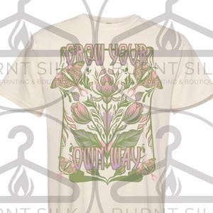 May include: Cream-colored t-shirt featuring a floral design with the text "Grow Your Own Way." The design includes pink and green flowers, leaves, and butterflies within a green rectangular frame. The shirt is a short-sleeved crew neck.