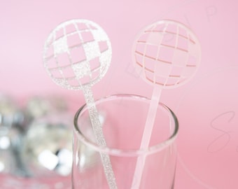 May include: Two disco ball drink stirrers in a champagne flute. One stirrer has a silver glitter handle and a pink and silver disco ball top. The other stirrer has a pink handle and a pink disco ball top. The background is pink.