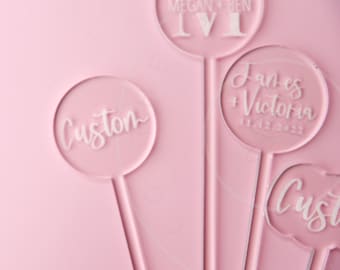 May include: Four clear acrylic circular cake toppers with white engraved text. One reads Megan + Ben, another James + Victoria 11.12.2022, and two read Custom. The toppers are arranged on a solid light pink background surface.
