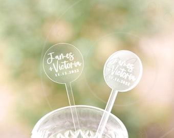May include: Two clear acrylic circular drink stirrers featuring white printed text that reads James + Victoria and the date 11.12.2022. The stirrers have a long, thin stem and a round top, designed for use in glassware for events.