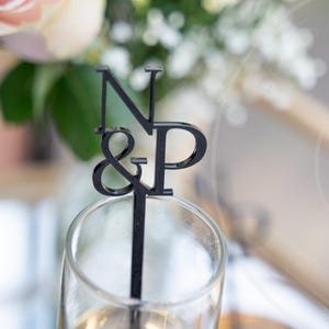 May include: A black acrylic cake topper with the letters "N & P" in a stylized font, inserted into a champagne glass. The background includes a bouquet of white flowers and a pink rose, suggesting a wedding or celebration theme.