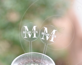 May include: Two clear circular acrylic drink stirrers featuring a white printed design. Each stirrer displays a large letter M with the text MEGAN + BEN printed across the center in a clean, sans-serif font. The stirrers have long, thin stems.
