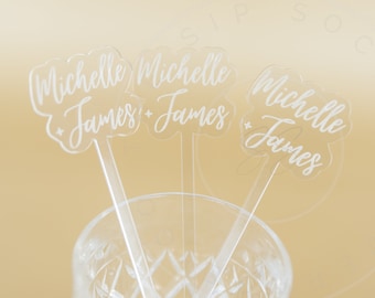 May include: Three clear acrylic drink stirrers with a scalloped top edge. Each stirrer features the white printed text Michelle + James in a cursive script font. The stirrers are positioned upright inside a clear glass container.