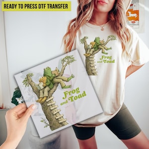 May include: A cream-colored t-shirt and a canvas featuring a whimsical illustration of Frog and Toad reading in a tree. The design includes the text "Frog and Toad" in green, and the words "READY TO PRESS DTF TRANSFER" in yellow.
