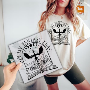 May include: A white t-shirt with a black graphic design of a dragon flying out of an open book. The graphic also includes crescent moons and the text "In My Fantasy Era."