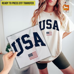 May include: Cream-colored t-shirt and a white square featuring the word "USA" in navy blue, with a small American flag. The t-shirt also has the word "USA" and a flag. The words "READY TO PRESS DTF TRANSFER" are at the top.