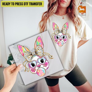 May include: A white t-shirt with a colorful cartoon bunny wearing heart-shaped sunglasses and a bow, blowing a bubble. The bunny has pink ears, a white face, and a pink nose. The bunny is surrounded by a gold glitter effect. The text "READY TO PRESS DTF TRANSFER" is at the top of the image.