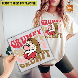 May include: A white t-shirt with a red, pink, and brown graphic design that says "Grumpy" multiple times. A cartoon image of Grumpy from Snow White is featured in the design.