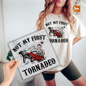 May include: A white t-shirt with the text "Not My First Tornado" and a cartoon image of a person riding in a red car through a tornado. The shirt is being held by a person's hand.