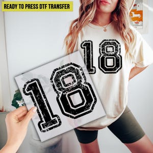 May include: A white canvas print featuring the number "18" in a distressed black font. The same number is printed on a cream-colored t-shirt. The text "READY TO PRESS DTF TRANSFER" is in yellow.