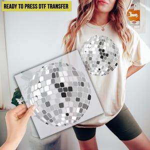 May include: A beige t-shirt with a disco ball graphic. A square print with a disco ball design is held in front of the shirt. The text "READY TO PRESS DTF TRANSFER" is in a yellow rectangle at the top.