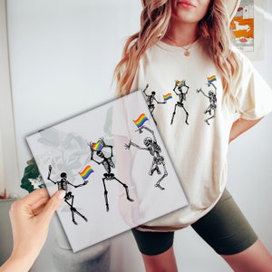 May include: A white t-shirt with a graphic of skeletons holding rainbow flags and hearts. The skeletons are dancing and smiling. The shirt is a great way to show your pride.