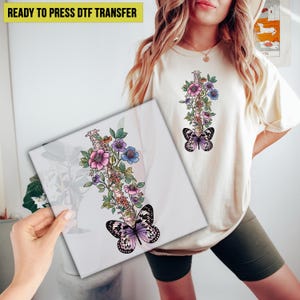 May include: A cream-colored t-shirt and a white square canvas featuring a floral and butterfly design. The design includes a spine-like structure adorned with pink, purple, and blue flowers, and a butterfly. The text "READY TO PRESS DTF TRANSFER" is at the top.