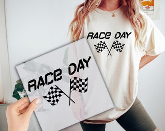 Op de afbeelding: Een wit T-shirt met de tekst "RACE DAY" en een zwart-wit geblokte vlag afbeelding. Het shirt wordt vastgehouden door een persoon.