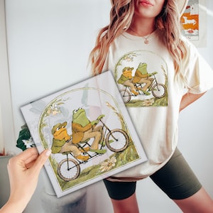 May include: A white t-shirt with a green and brown illustration of two frogs riding a bicycle. The frogs are wearing suits and hats. The illustration is surrounded by a green border.