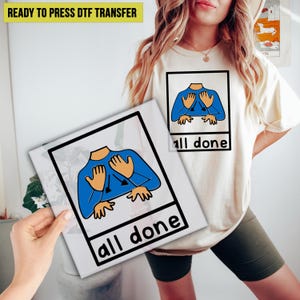 May include: A cream-colored t-shirt with a graphic of a blue shirt with hands and the words "all done" in black. A ready-to-press DTF transfer is held in front of the shirt, featuring the same design. The image is of a person wearing the shirt.