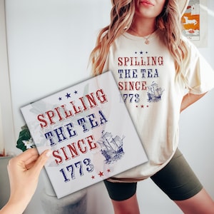 May include: White t-shirt with red, white, and blue text that reads "Spilling the Tea Since 1773" and a blue and white illustration of a ship. The shirt is worn by a person with long blonde hair and is paired with black shorts.