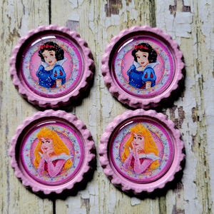 Peut inclure: Quatre aimants en forme de bouchon de bouteille rose et blanc avec des images de Blanche-Neige et de la Belle au bois dormant. Les aimants ont un fond rose avec une bordure blanche et un revêtement époxy transparent.