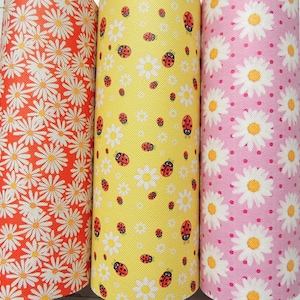 May include: Three rolls of fabric featuring a daisy and ladybug design. The fabric on the left is orange with white daisies. The middle fabric is yellow with white daisies and red ladybugs. The fabric on the right is pink with white daisies and pink polka dots.