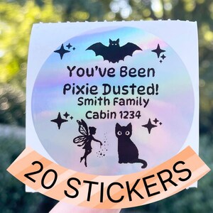 May include: A round sticker with a holographic background featuring a bat, stars, a fairy, and a black cat. The text reads "You've Been Pixie Dusted! Smith Family Cabin 1234." A banner at the bottom says "20 STICKERS."