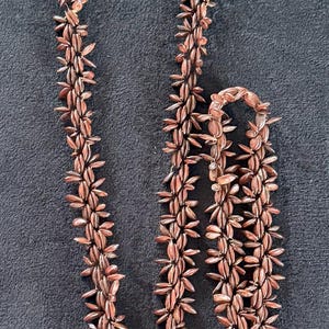 May include: A necklace composed of numerous small, copper-colored, leaf-shaped elements. The necklace is arranged in a repeating pattern, creating a textured and visually interesting design. The necklace is displayed against a dark background.