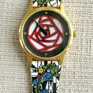 May include: A gold-tone wristwatch with a black face and a red rose design. The watch band is white with a black and blue floral pattern.