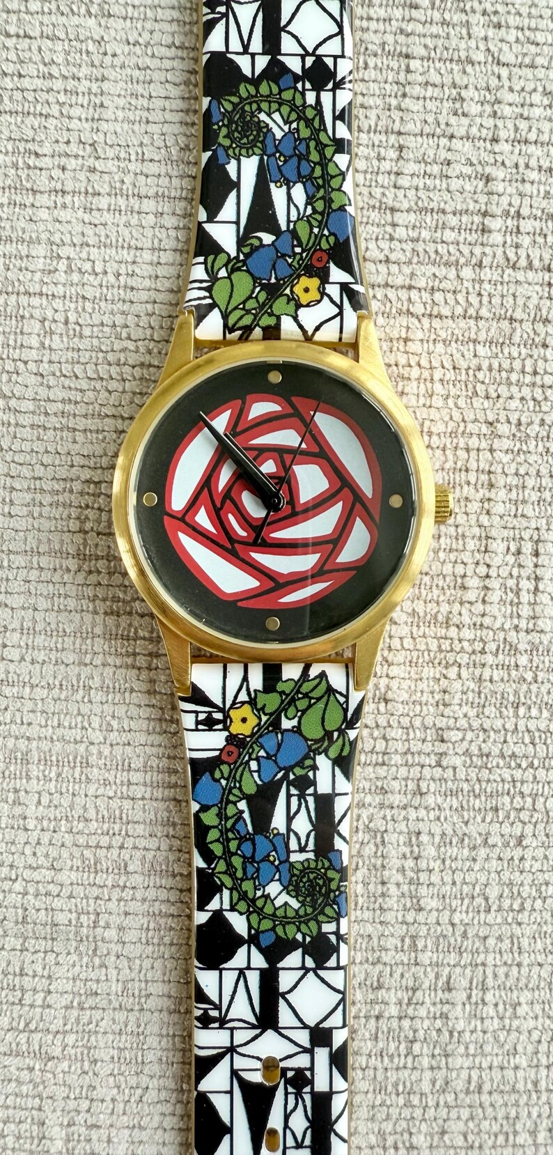 May include: A gold-tone wristwatch with a black face and a red rose design. The watch band is white with a black and blue floral pattern.