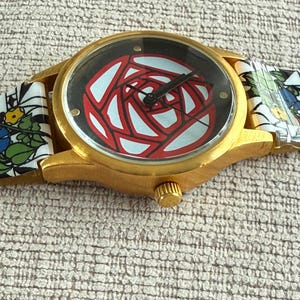 May include: A gold-tone wristwatch with a black face and a red and white stylized rose design. The watch has a floral patterned band.