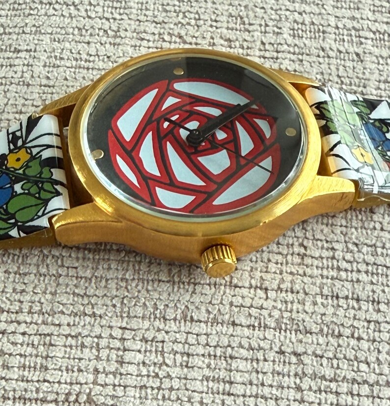 May include: A gold-tone wristwatch with a black face and a red and white stylized rose design. The watch has a floral patterned band.