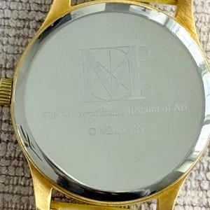 May include: Gold-tone watch with a round, silver-colored face. The back of the watch has an engraved logo for The Metropolitan Museum of Art.