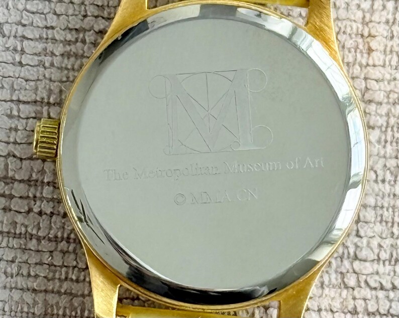 May include: Gold-tone watch with a round, silver-colored face. The back of the watch has an engraved logo for The Metropolitan Museum of Art.