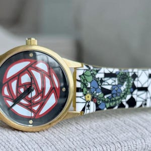 May include: A gold wristwatch with a white and black patterned band. The watch face features a red and white rose design.
