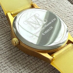 May include: Gold-tone wristwatch with a yellow rubber band. The back of the watch has an engraved logo for The Metropolitan Museum of Art and the copyright symbol with the letters "MMACN".