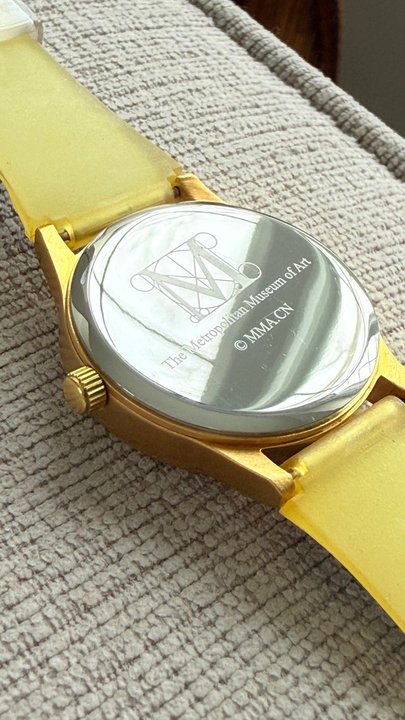 May include: Gold-tone wristwatch with a yellow rubber band. The back of the watch has an engraved logo for The Metropolitan Museum of Art and the copyright symbol with the letters "MMACN".