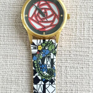 May include: A gold-tone wristwatch with a black face and a red rose design. The watch band is white with black geometric patterns and floral accents.