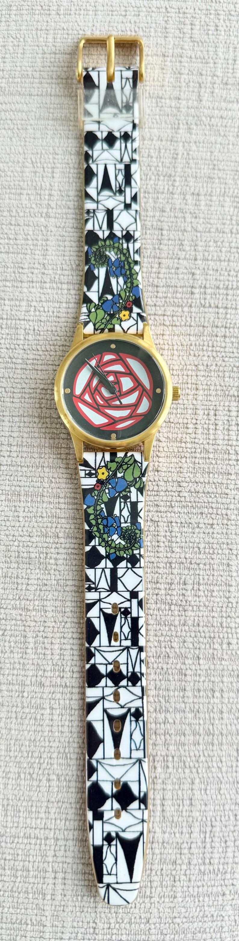 May include: A gold-tone wristwatch with a black face and a red rose design. The watch band is white with black geometric patterns and floral accents.